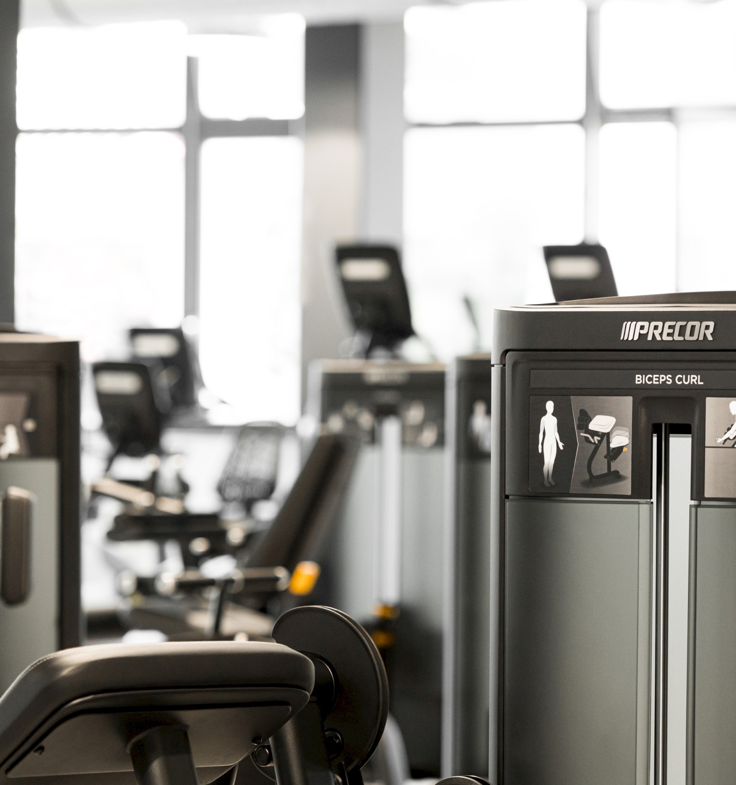 The image shows gym equipment, specifically weight machines, in a fitness center setting with natural light from large windows.