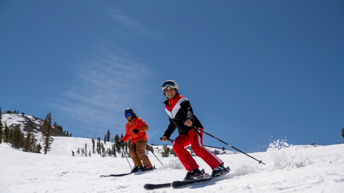 Two people skiing down a snowy slope under a clear blue sky, wearing colorful ski outfits and helmets for protection.