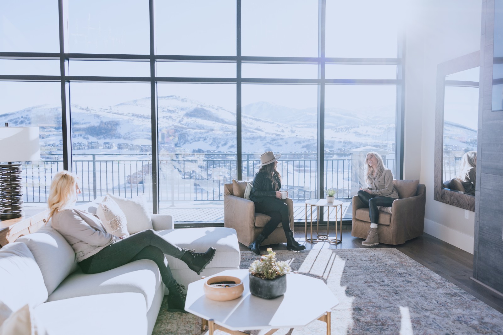 People are relaxing and chatting in a cozy living room with large windows showing snowy mountains outside.