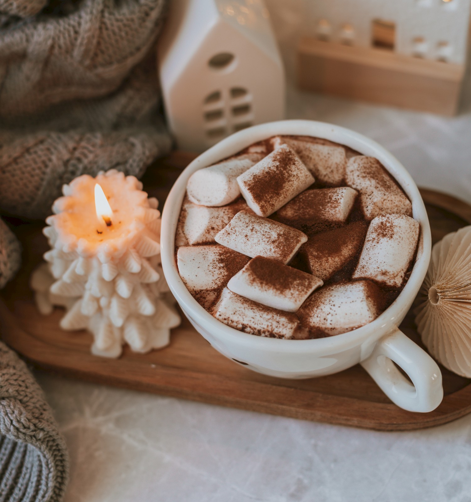 A cozy scene with hot chocolate topped with marshmallows, a lit candle, knitted fabric, and festive decorations on a wooden tray.