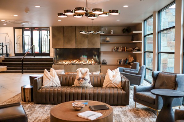 A cozy living room with a leather sofa, armchairs, fireplace, bookshelves, and large windows letting in natural light.