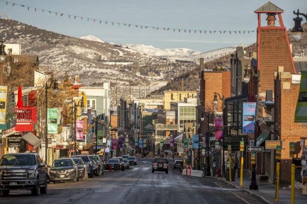 A picturesque street with parked cars, colorful buildings, and snow-covered mountains in the background.