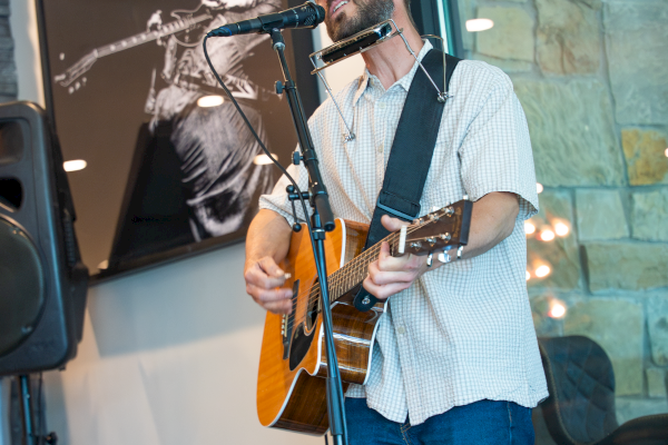A musician is performing indoors, playing guitar and singing into a microphone, with artwork and a TV in the background.