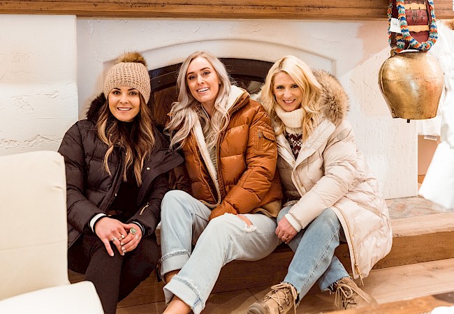 Three people in winter jackets sit on a hearth, surrounded by coats and winter decor, smiling.