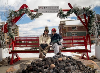 Two people sit on a red bench by a fire pit at Park City Canyons Village, with snow and buildings in the background.
