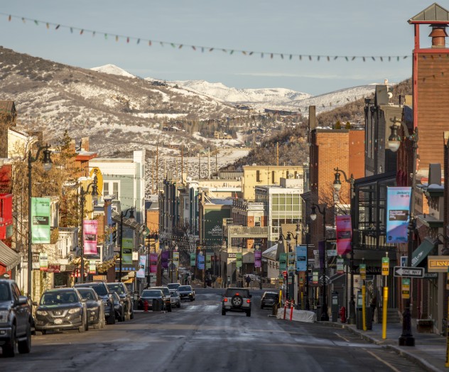 A scenic downtown street with colorful storefronts, parked cars, and snowy mountains in the background on a clear day.