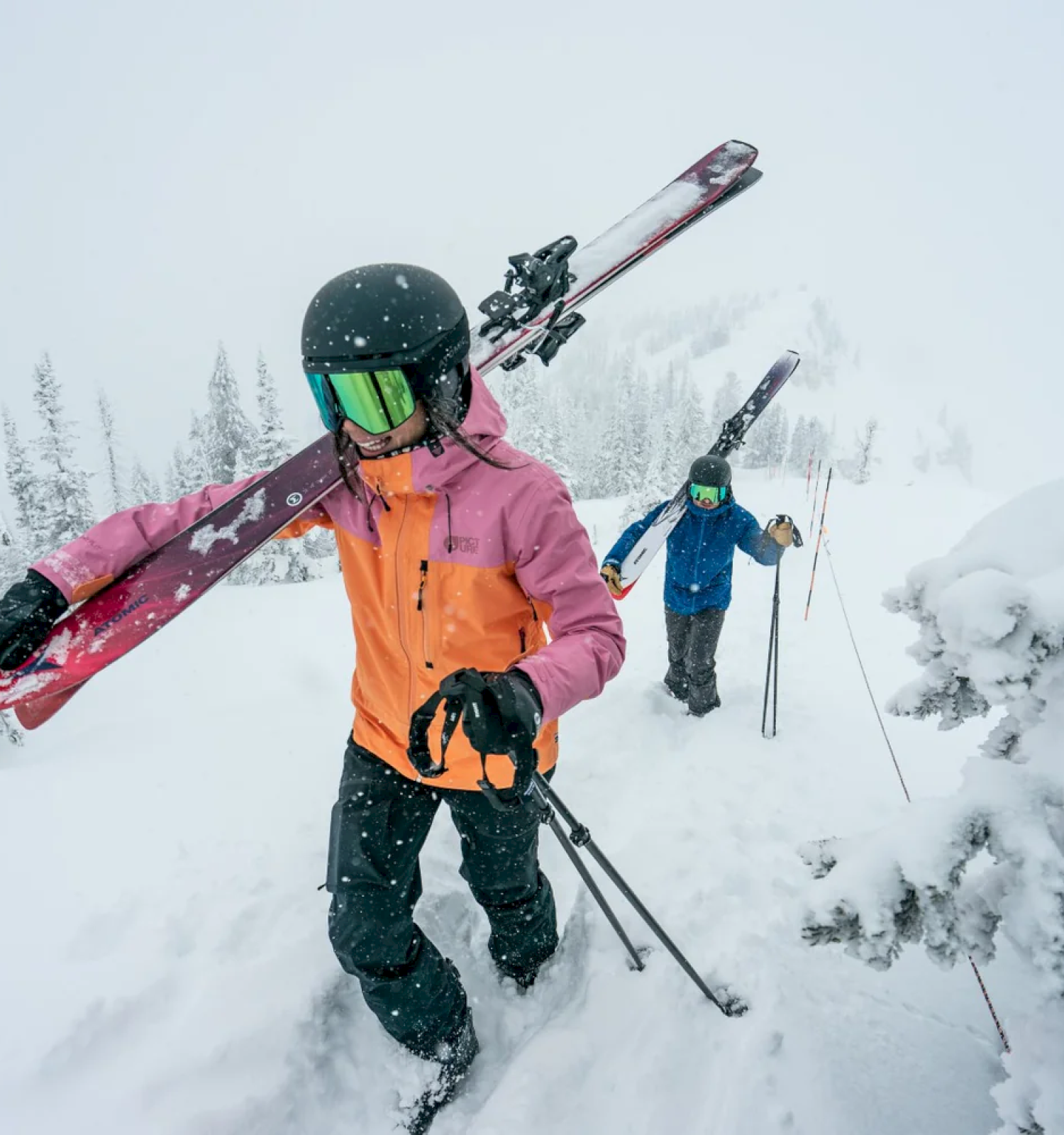 Two people hike through a snowy landscape, each carrying skis and poles, dressed warmly for winter conditions.