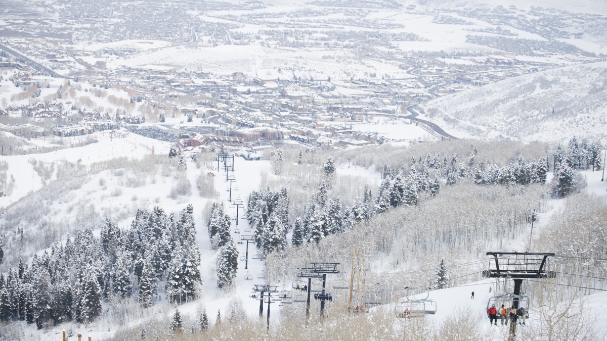 A snowy landscape with chairlifts moving above snow-covered trees, overlooking a distant town or city in the valley.