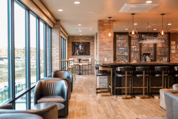 The image shows a modern lounge bar with brick walls, a row of bar stools, leather chairs, and large windows offering scenic views.