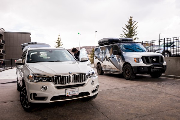 A white BMW SUV and a van with a mountain design are parked on a concrete surface near a building. A person is near the SUV's trunk.