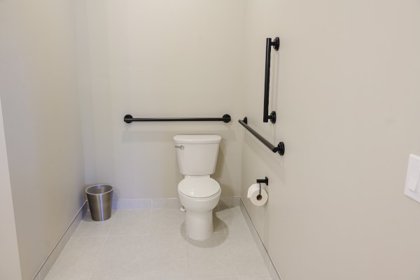 The image shows a restroom with a toilet, grab bars, a toilet paper holder, and a trash can. The walls and floor are light-colored.