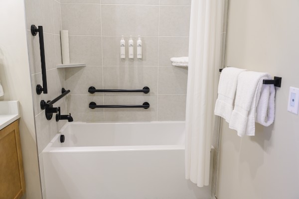 The image shows a bathroom with a bathtub, wall-mounted soap dispensers, a towel on a rail, and safety grab bars.