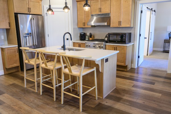 A modern kitchen with wooden cabinets, a central island with stools, pendant lights, stainless steel appliances, and a sleek faucet.