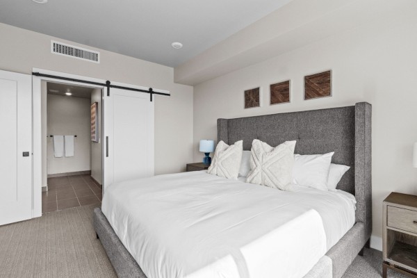 A modern bedroom features a large bed with white bedding, a gray headboard, pillows, wooden side tables, and a sliding barn door.