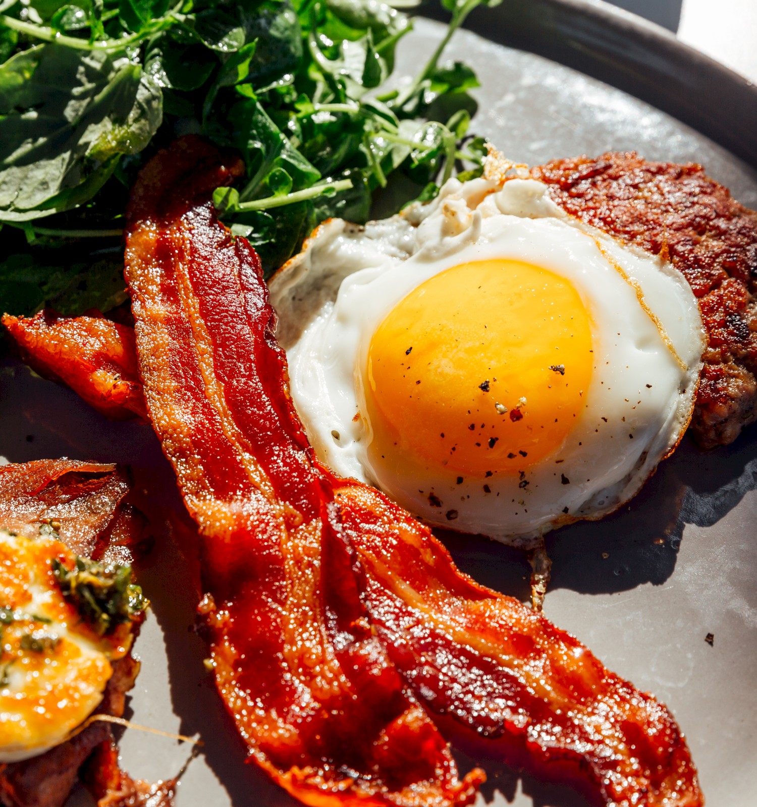 A breakfast plate with bacon, a sunny-side-up egg, greens, and a hash brown patty on a round dish.