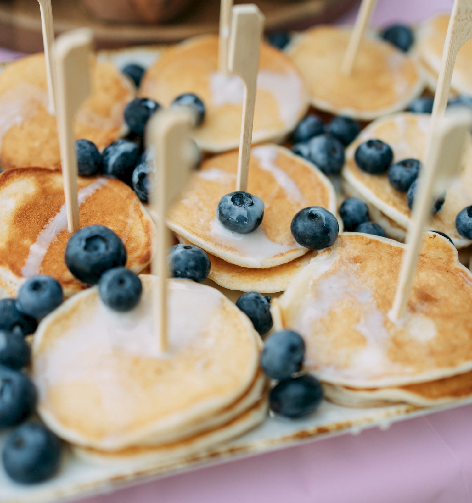 Mini pancakes topped with blueberries and wooden sticks are arranged on a tray.