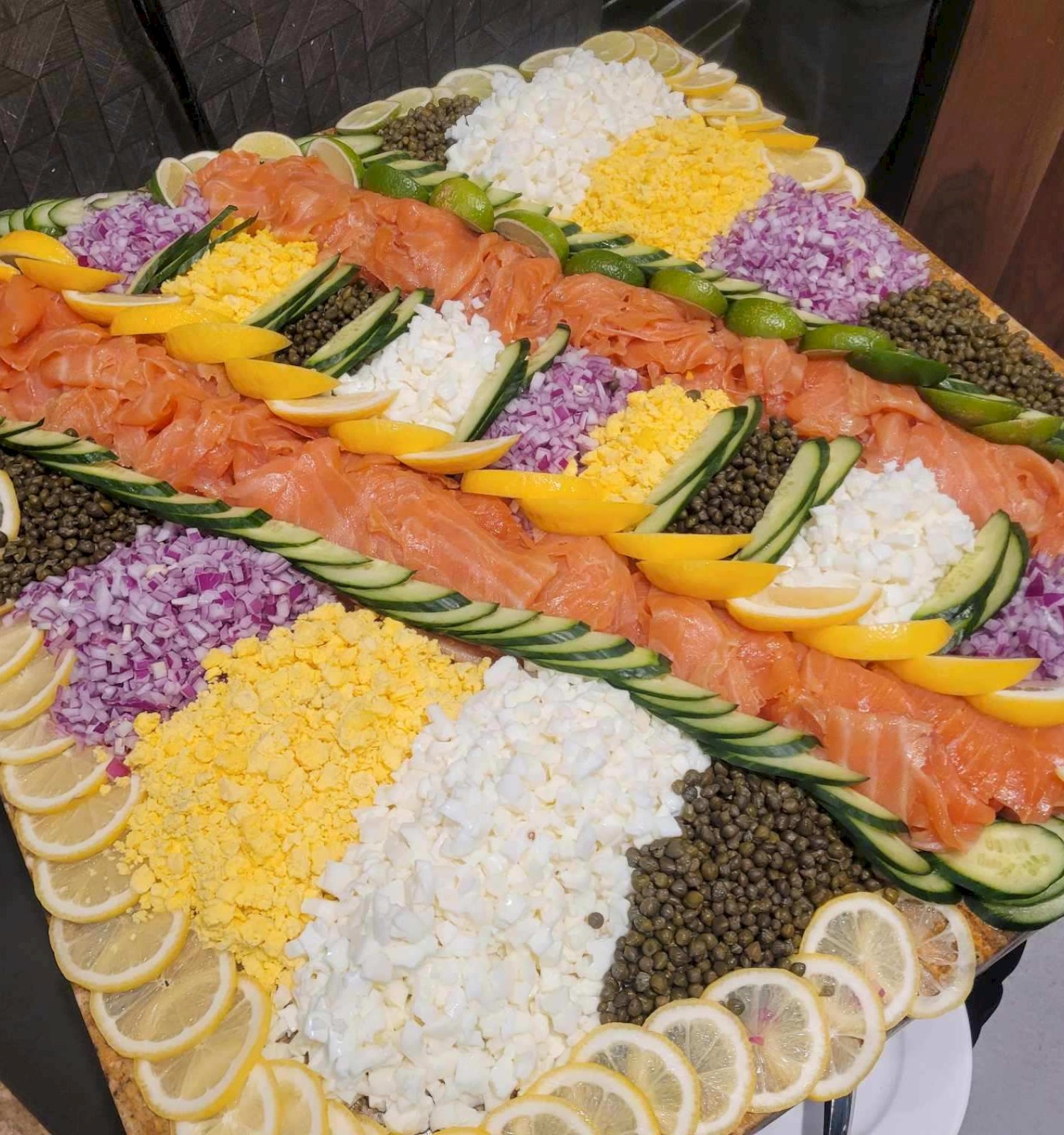 A platter with smoked salmon, capers, lemon slices, cucumber, diced red onion, chopped egg, and cream cheese arranged decoratively on a board.