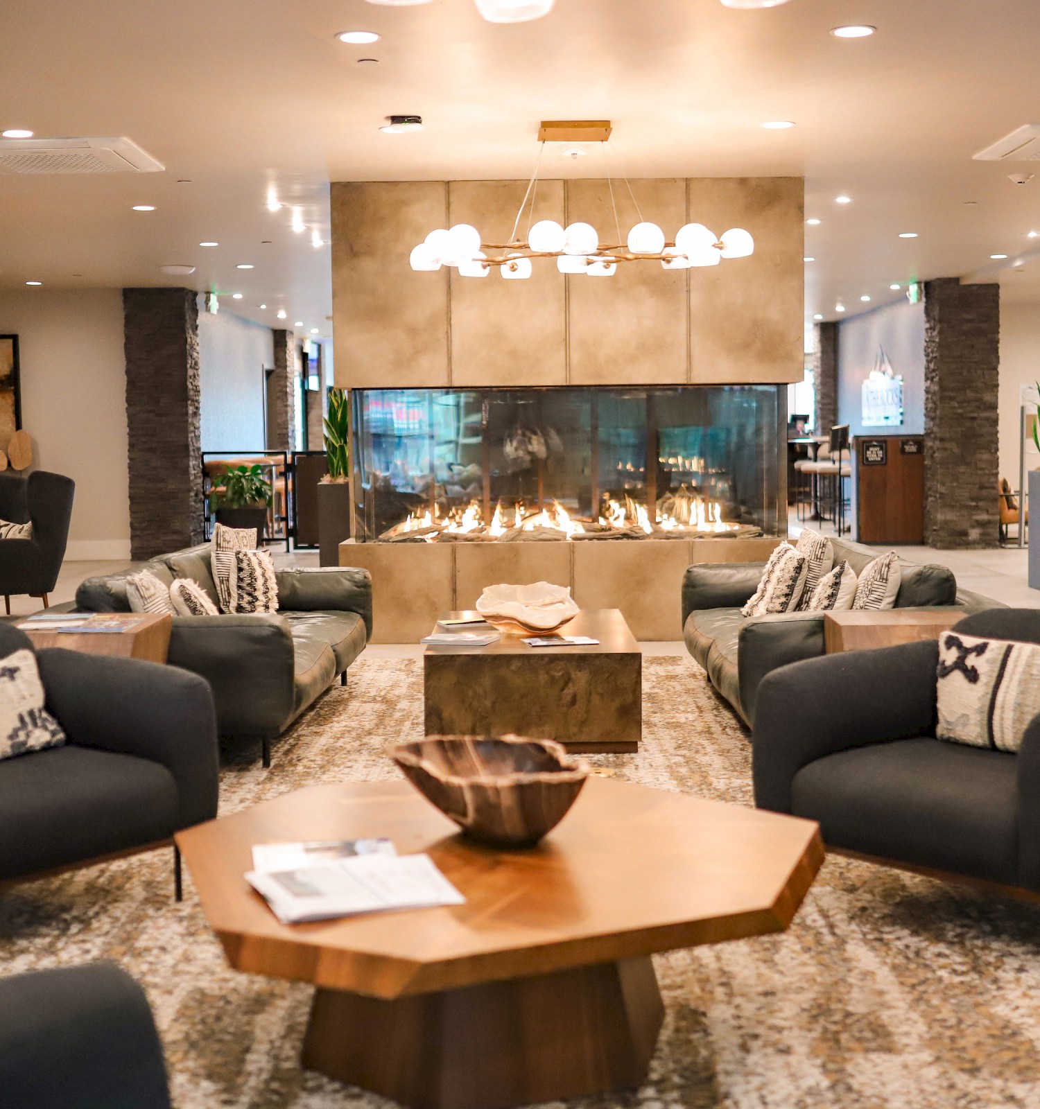 A cozy lounge features dark sofas, a wooden table, and a large fireplace surrounded by warm lighting and modern decor accents.