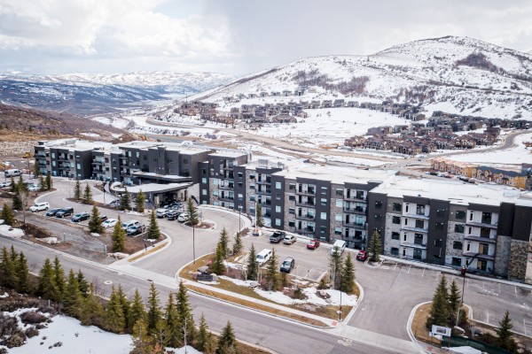 Aerial view of a large building with parking, surrounded by snowy mountains and trees, under a cloudy sky.