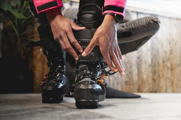 A person is adjusting ski boots, ensuring a secure fit, with one knee on the ground and a hand on each boot clasp for stability.