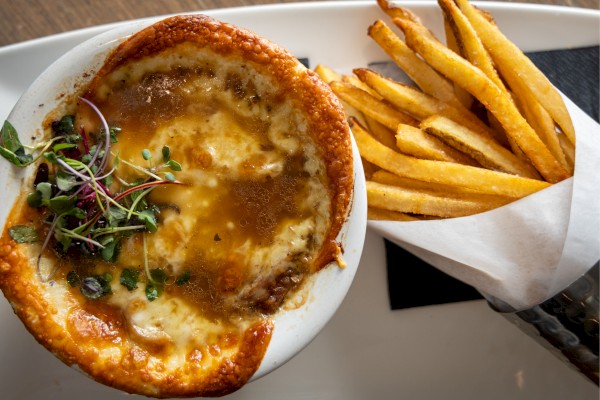A bowl of French onion soup topped with melted cheese and herbs is next to a serving of golden fries in a paper cone.