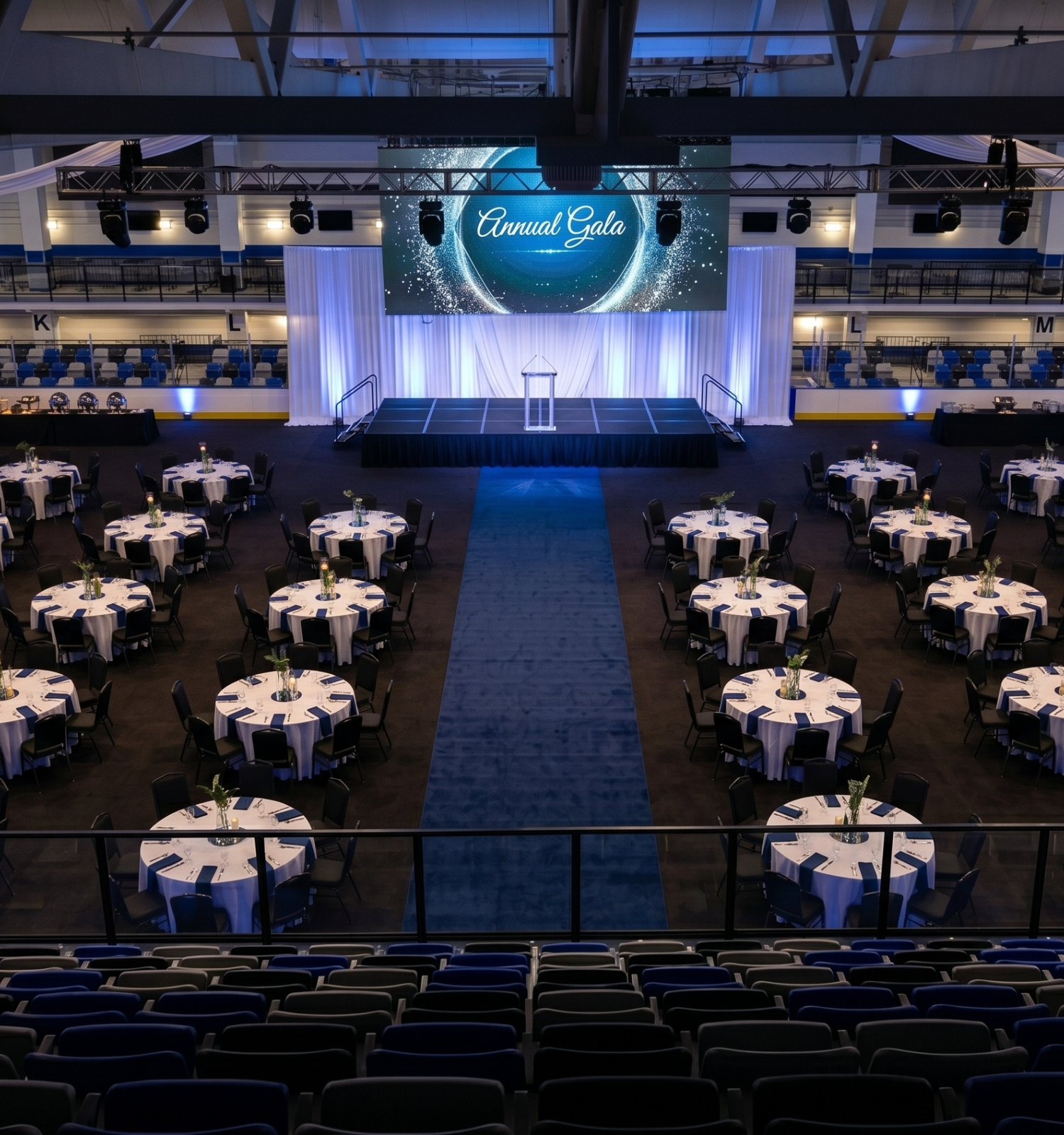 The image shows an empty banquet hall set up for an annual gala with round tables, a stage at the front, and a large screen displaying 