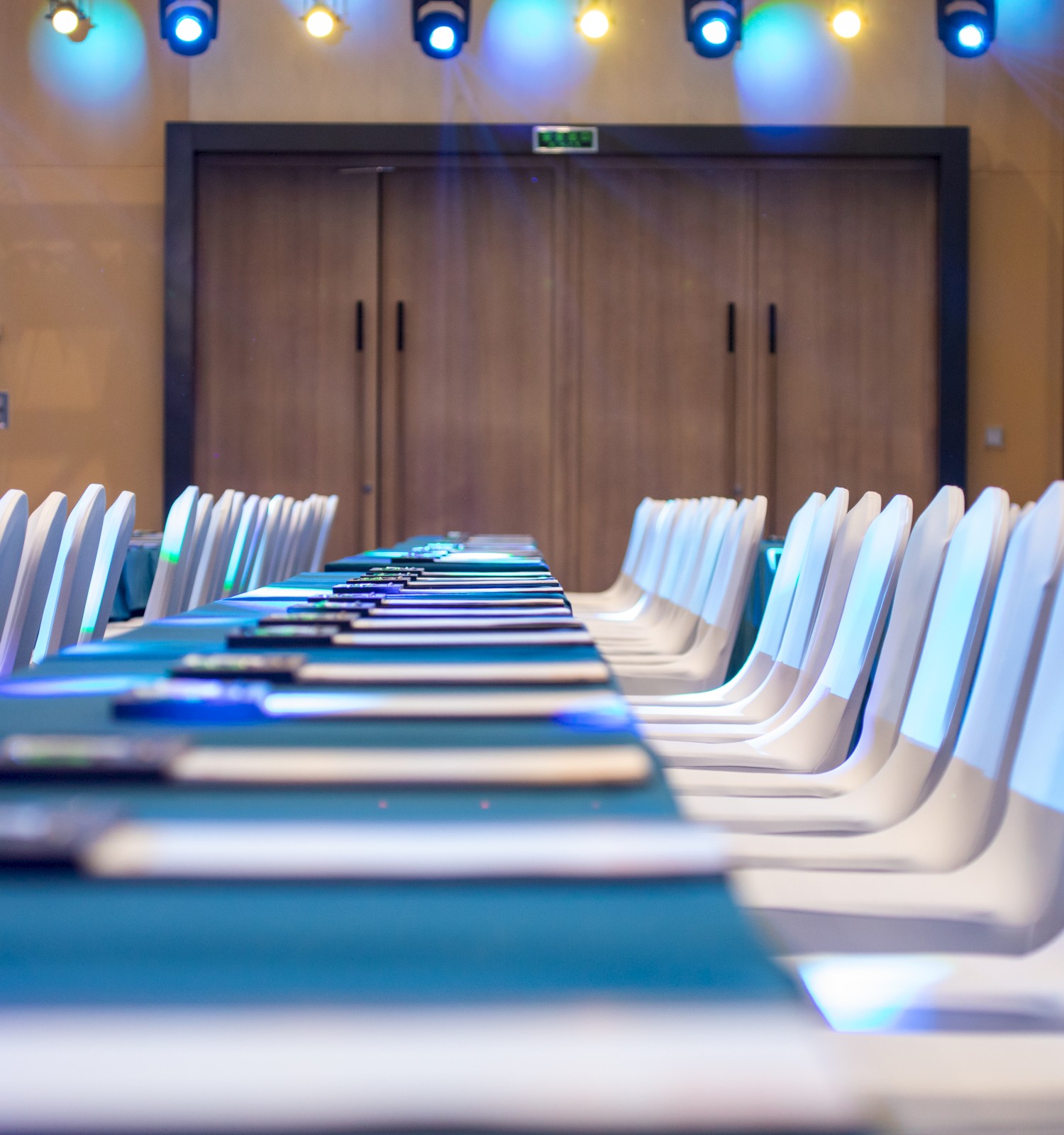 A long conference table with white chairs is set up in a room with blue lighting and closed double doors in the background.