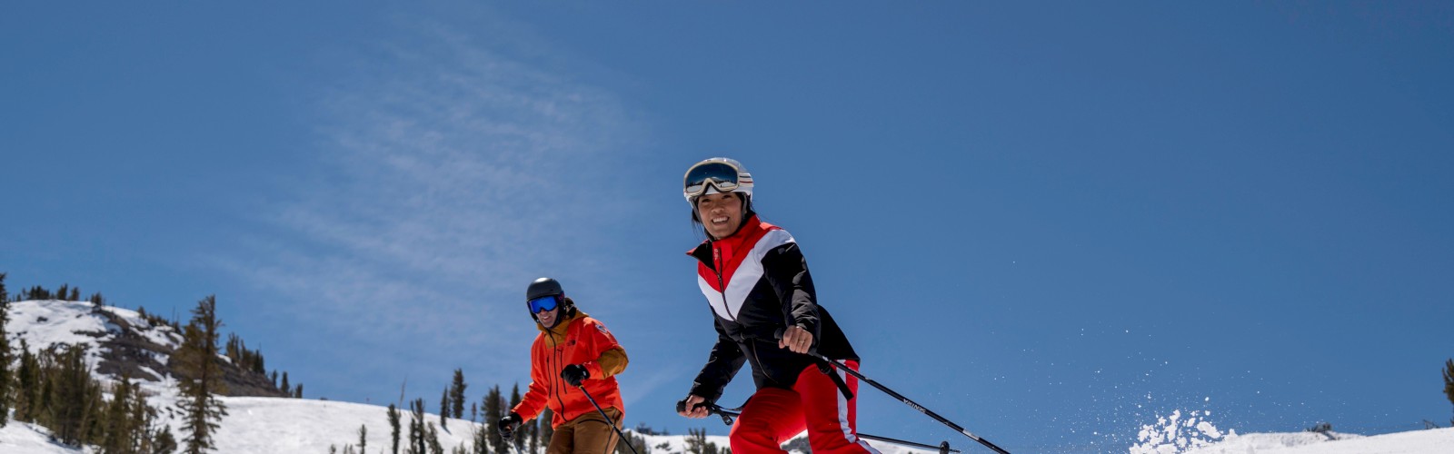 Two people skiing on a snowy slope under a clear blue sky, surrounded by a few scattered trees in the background.