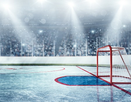 An empty ice hockey rink under bright lights, featuring a goalpost and an audience in the background.