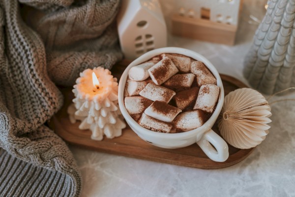 A cozy scene with hot chocolate topped with marshmallows, a lit candle, knitted blanket, and decorative items placed on a wooden tray.