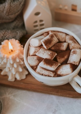 A cozy scene with a cup of hot chocolate topped with marshmallows, a lit candle, and warm decor on a wooden tray.
