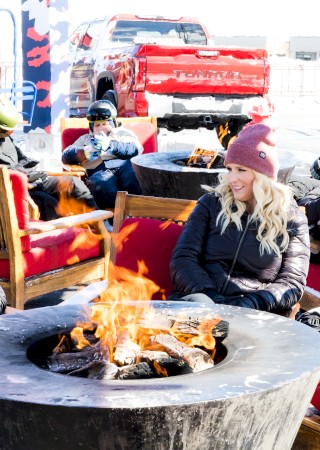 People sitting around a fire pit in a sunlit outdoor lounge, wearing winter clothes and beanies, chatting and relaxing.