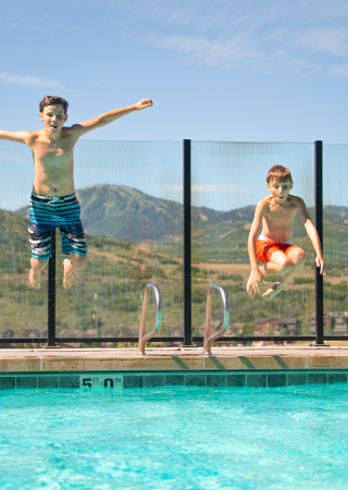 Two boys are jumping into a swimming pool from the deck on a sunny day with mountains in the background.