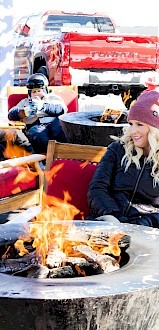 A group of friends sits around a lit fire pit on a sunny outdoor deck, wearing jackets and beanies near a red truck and lounge seating.