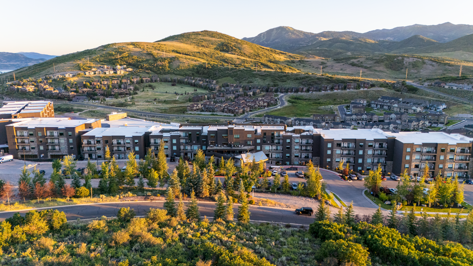 A modern apartment complex sits at the foothill of green, rocky hills with a winding road and trees in the foreground.