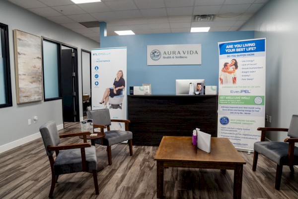 A modern reception area with a dark wooden desk, several chairs, a central table, and informational banners about a health or wellness clinic.