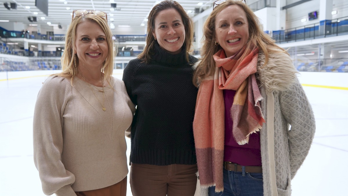 Three women pose together on an ice rink, smiling, inside a modern arena with bright lights and empty seating in the background.