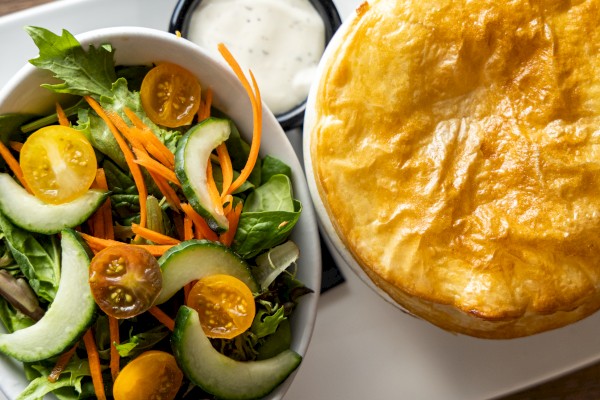 A fresh garden salad with cucumber, cherry tomatoes, carrot ribbons, greens, and herbs next to a golden-brown pie or casserole on the right.