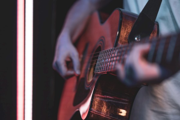 A person plays an acoustic guitar, fingers strumming near the sound hole, close-up with soft lighting and a vertical neon edge nearby.