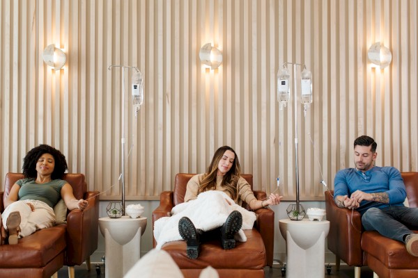 Three people sit in a medical salon or lounge with IV stands beside them, relaxing on orange chairs under warm wall lights.
