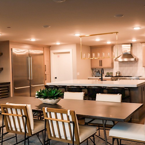 A modern open-concept kitchen and dining area with a large island, stainless steel appliances, and a long dining table plus wooden chairs, warmly lit.