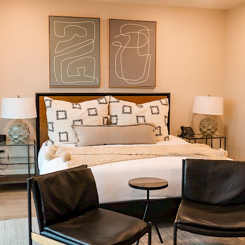 A stylish bedroom with a large bed, two nightstands, matching lamps, abstract art above, and two black chairs with a small table in front.