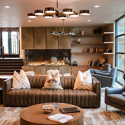 A modern, cozy hotel lobby with a central sofa seating area, shelving, warm lighting, and large windows, inviting and stylish.