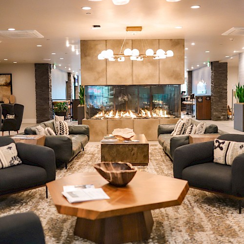 Cozy hotel lobby with dark couches, central wooden coffee table, warm lighting, fireplace, plants, and a modern, inviting seating area.