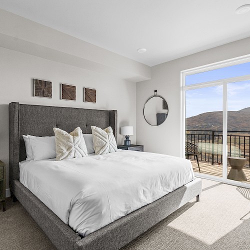 A bright bedroom with a large bed, gray headboard, two nightstands, lamps, and a sliding glass door opening to a balcony with hills in the background.