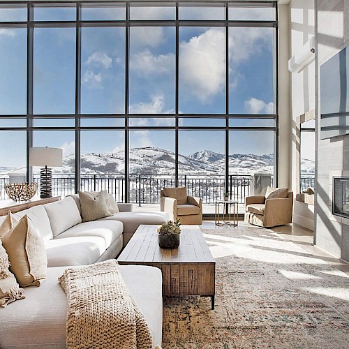Luxurious living room with oversized windows, snow-capped mountains outside, white sectional sofa, neutral cushions, woven ottoman, and modern decor.