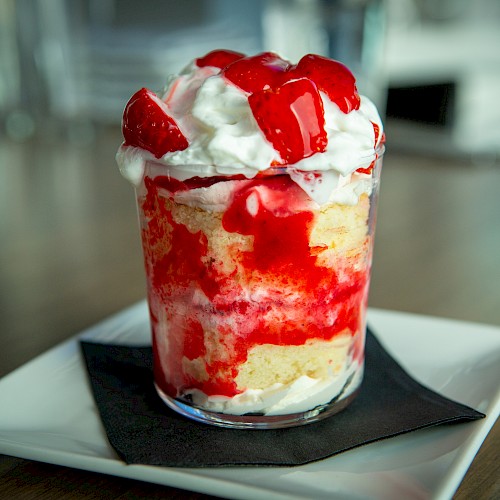 A tall glass dessert with vanilla ice cream, whipped cream, and strawberry sauce drizzled over a bed of crumbs, served on a white plate.