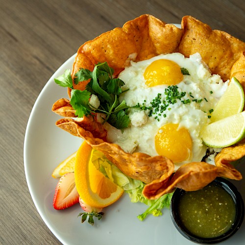 Delicious breakfast tostada bowl with fried eggs, pico de gallo, avocado, lime wedges, greens, tortilla bowl, and a side of green salsa.