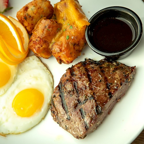 A plate with sunny-side eggs, crispy fried potatoes, steak slices, orange wedges, and a small cup of dipping sauce. End.