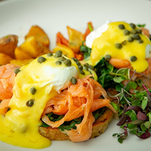 Two poached eggs over smoked salmon and toasted bread, topped with hollandaise, served with crispy potatoes and a greens garnish.